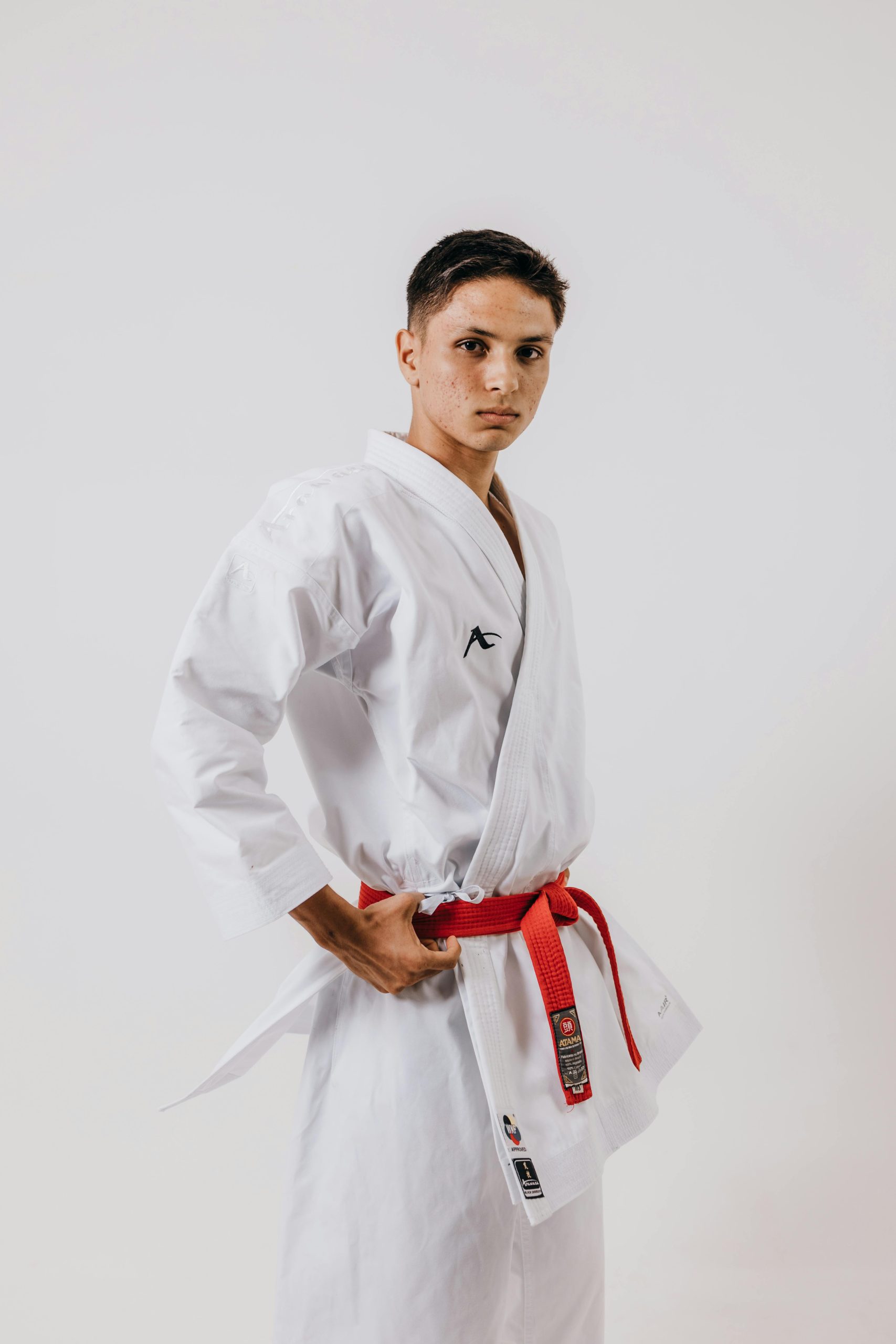 A teenage karate student posing confidently in a red belt with arms akimbo on a white background.